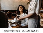 Young smiling african woman using smartphone while paying restaurant bill with contactless payment during dinner party with friends
