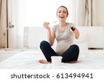 Cheerful pregnant young woman with ice cream laughing and watching TV on bed at home