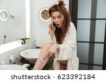 Astonished shocked young woman in bathrobe shaving legs and talking on mobile phone in bathroom
