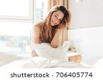 Positive woman with pillow sitting in bed and smiling to camera at home