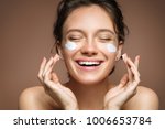 Laughing girl applying moisturizing cream on her face. Photo of young girl with flawless skin on brown background. Skin care and beauty concept