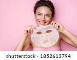 Smiling woman holds facial sheet mask. Photo of attractive woman with perfect skin on pink background. Beauty & Skin care concept