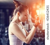 Small photo of Young slim woman doing pushdown on cable machine in gym in sunset beams. Athletic girl training triceps in fitness center