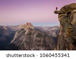 A fearless male hiker is standing on an overhanging rock at Glacier Point enjoying the breathtaking view towards famous Half Dome in beautiful post sunset twilight, Yosemite National Park, California