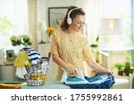happy modern middle age woman in yellow dress with washed clothes basket ironing on ironing board while listening to the music with headphones in the modern house in sunny day.
