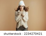 Hello winter. smiling stylish woman in beige sweater, mittens and hat on beige background blowing snow.