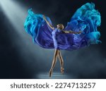 Ballerina in Purple Chiffon Dress on Stage Light Beam. Ballet Dancer in Silk Fantasy Blue Gown. Woman dancing with flying Cyan Fabric as Wings over Dark Background