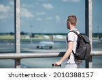 Traveler watching airplanes at airport. Rear view of passenger with backpack looking through window of terminal building.