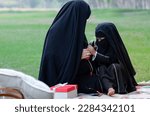 Muslim mother and daughter wearing black niqab spending weekends relaxing in the park, sitting on picnic blankets in the park, little girl using headphones