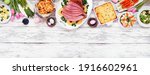Small photo of Traditional Easter ham dinner. Overhead view top border on a white wood banner background with copy space. Ham, scalloped potatoes, vegetables, eggs, hot cross buns and carrot cake.