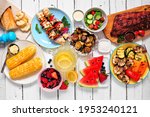 Summer BBQ or picnic food concept. Assortment of grilled meats, vegetables, fruits, salad and potatoes. Top down view table scene with a white wood background.