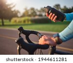 Hydrate your body. Close up of hands of professional cyclist holding water bottle, standing with his bike in park on a sunny day
