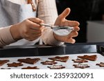 culinary, cooking and christmas concept - close up of hands sifting powdered sugar through sieve to raw gingerbread cookies on baking tray
