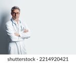 Small photo of Portrait of trustworthy older smart doctor with gray hair wearing glasses and white lab coat standing against white wall, smiling. Copy space.