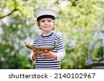 Small photo of Happy little kid boy in sailor capitain hat and uniform playing with sailor boat ship. Smiling preschool child dreaming and having fun. Education, profession, dream concept