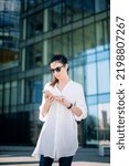Small photo of Careworn woman using smartphone and walking outdoors in city. Confident female wearing white shirt and jeans and text messaging.