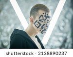 Small photo of Modern technologies of human personification. Portrait of a young man with a QR-code painted on his face, who stands in front of futuristic neon lamps and looks at the camera unfriendly and aloof.