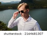 A purposeful young man in a white shirt and black sunglasses stands against a lake and mountain landscape. Business concept. Vacation and travel.