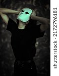 Small photo of A girl in black clothes with a mask on her face suffering and holding her head with her hands. Dark grunge background. Human roles. Hypocrisy. Mental disorders.