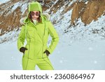 Winter sport in the mountains. Portrait of a fashionable girl posing in green ski suit against a snowy winter landscape. Winter fashion. 