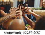 Small photo of A diverse group of friends, in their forties, with various ethnicities and tattoos, gather around a table for dinner. They enthusiastically join hands in a high-five, blurred faces and focus on hands