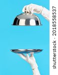 Small photo of Elegant waiter's hands in white gloves holding silver tray and cloche on blue background. Restaurant, horeca, first class service concept