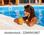 Small photo of Attractive young woman leaning on the edge of a swimming pool, relaxing while on summer vacation, sunbathing and drinking cocktails