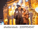Beautiful young couple in love having fun celebrating New Year on city streets or square, holding sparklers for midnight countdown with Christmas lights in background
