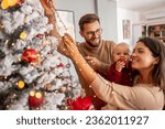 Young parents having fun decorating Christmas tree with their cute little baby girl, placing Christmas lights on it while decorating home for winter holiday season