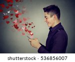 Side profile happy man sending love sms text message on mobile phone with red hearts flying away from screen isolated on gray wall background. Positive human emotions