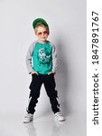 Small photo of Cool impish frolic blond kid boy buddy in cap, sunglasses grey sweatshirt with dinosaur print, black sport pants with stripes and white sneakers stands holding hands in his pockets
