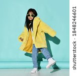 Small photo of Frolic smiling asian kid girl in stylish yellow furry coat, jeans, sneakers and sunglasses dances stands with her foot up over blue wall background. Trendy children fashion, stylish outfit concept