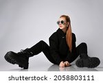 Cool trendy rich red-haired woman rock star, biker in modern black hoodie, pants and massive brutal army shoes sits on floor with legs wide apart and looks aside at copy space
