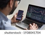 Small photo of Business man trader investor analyst using mobile phone app analytics for cryptocurrency financial market analysis, trading data index chart graph on smartphone and laptop screen. Over shoulder view