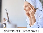Small photo of Gorgeous mid age older adult 50 years old blonde woman wears bathrobe in bathroom applying nourishing antiage face skin care cream treatment, looking at mirror doing daily morning beauty routine.