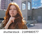 Hipster teen stylish cool redhead fashion girl model showing shh sign asking to keep secret, be hush silent or privacy silence standing in big city urban location. Headshot portrait