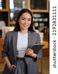 Small photo of Young smiling successful professional leader Asian business woman, female executive manager, saleswoman wearing suit holding digital tablet standing in office looking at camera, vertical portrait.