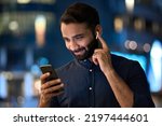 Small photo of Smiling bearded indian man wearing earbud holding phone having video call at night. Eastern businessman in earphone using smartphone listening music in app tech on cellphone watching videos online.