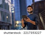 Smiling happy young eastern indian business man professional manager standing outdoor on street holding using digital tablet online fintech in night city with urban lights looking at camera, portrait.
