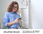 Small photo of Smiling happy young business woman manager holding smartphone working standing in office using mobile cell phone, checking financial banking apps, browsing online on cellphone typing on cellular.