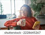 Small photo of Young happy woman using cell phone drinking coffee relaxing at home. Smiling pretty lady holding smartphone enjoying mobile shopping, daydreaming checking news on smartphone in cozy morning.