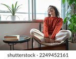 Young relaxed smiling pretty woman relaxing sitting on chair at home. Happy positive beautiful lady feeling joy enjoying wellbeing and lounge chilling near window in modern cozy apartment interior.