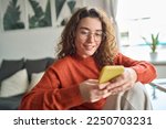 Young smiling woman wearing glasses holding smartphone using cellphone modern technology, looking at mobile, checking cell phone apps, texting, browsing internet for shopping sitting at home.