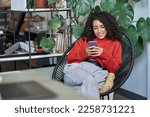 Small photo of Young smiling business woman looking at smartphone using cellphone mobile cellular, professional female worker or student typing on mobile cell phone sitting in chair with green plants around.
