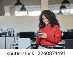 Smiling young professional latin business woman, happy lady corporate leader holding cellular phone working standing in modern office using mobile apps cellphone technology device looking at cell.