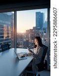 Small photo of Young busy Asian business woman using laptop and mobile cell phone tech at night in dark office. Professional businesswoman holding smartphone, working on cellphone with evening city view. Vertical