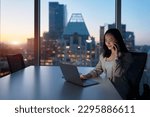 Young busy Asian business woman executive working on laptop making call at night in dark corporate office. Professional businesswoman manager talking to client using computer, city evening window view
