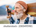 Small photo of Active cool bearded old hipster man standing in nature park holding skateboard wearing earbud. Mature traveler skater enjoying freedom spirit and extreme sports hobby listening music in earphones.