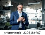 Happy middle-aged business man ceo wearing blue suit standing in office using cell phone. Older businessman professional executive holding mobile satisfied with enterprise solution management service.