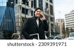 Small photo of Happy smiling young Asian business man professional holding cellphone walking on big city urban street making business call, talking on cellular phone standing in office downtown.